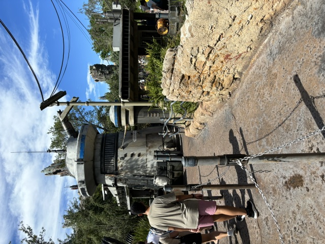 A metallic droid structure with cylindrical body and dome top at Star Wars: Galaxy's Edge, surrounded by rocky terrain and visitors