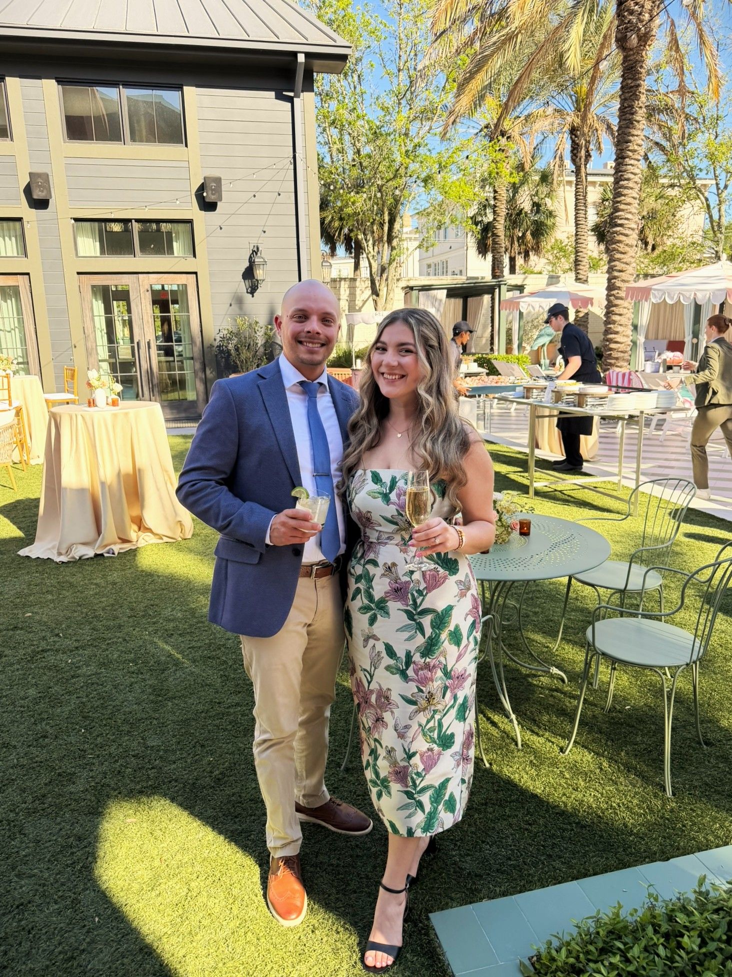Couple in formal attire smiling at outdoor garden reception with palm trees and catering tents