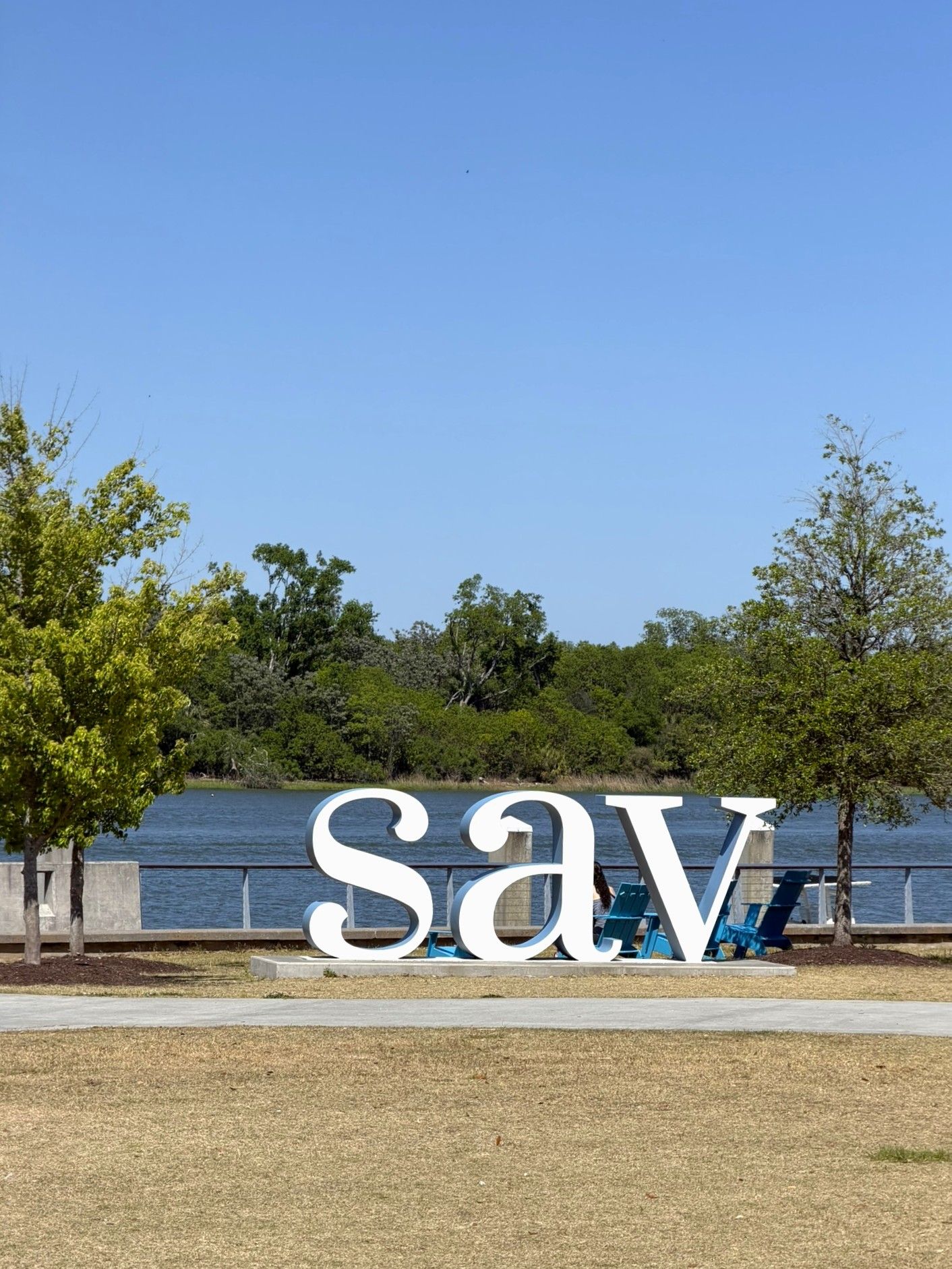 Large white 'SAV' letters on sandy ground with river and trees in background