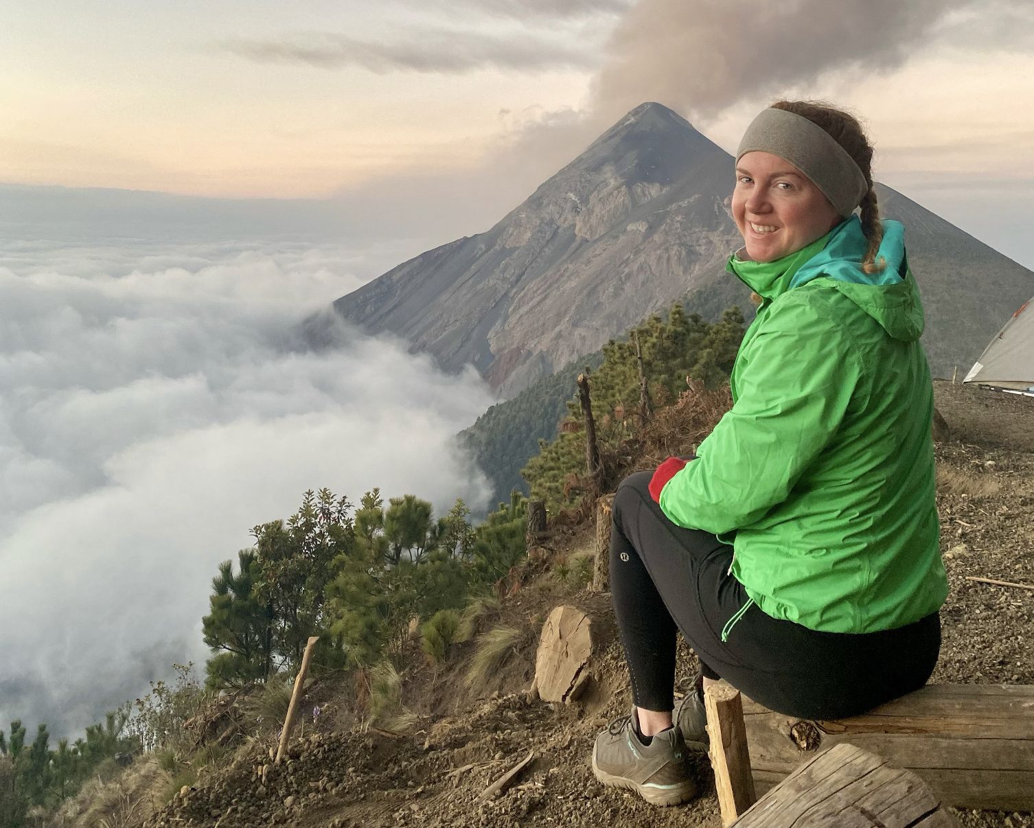 Autumn Fiske crouches on a wooden platform overlooking a volcanic cone shrouded in clouds, with forested slopes below in Guatemala