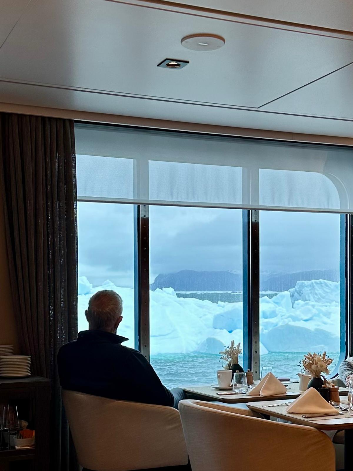 A man gazes out at icebergs from a ship's dining room with large windows overlooking the fjord