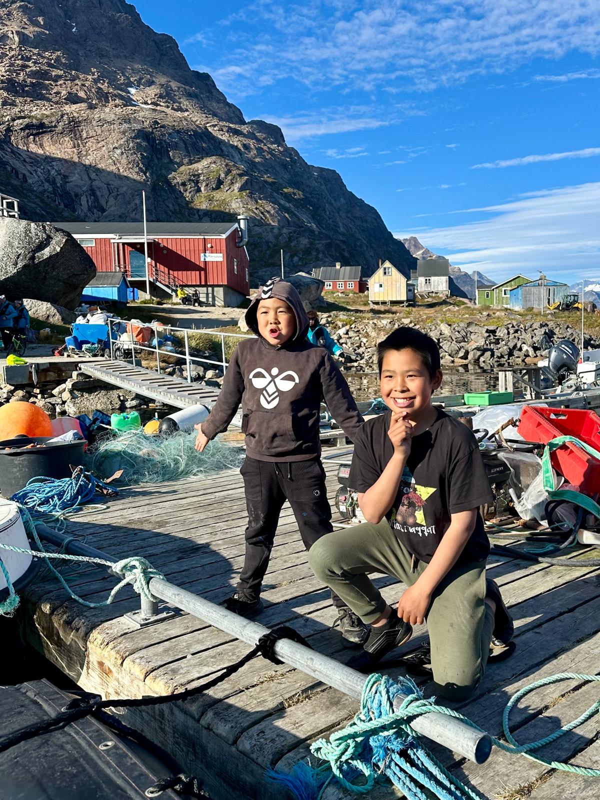 Two children stand on a wooden dock in a Greenlandic settlement with colorful houses and steep mountains behind