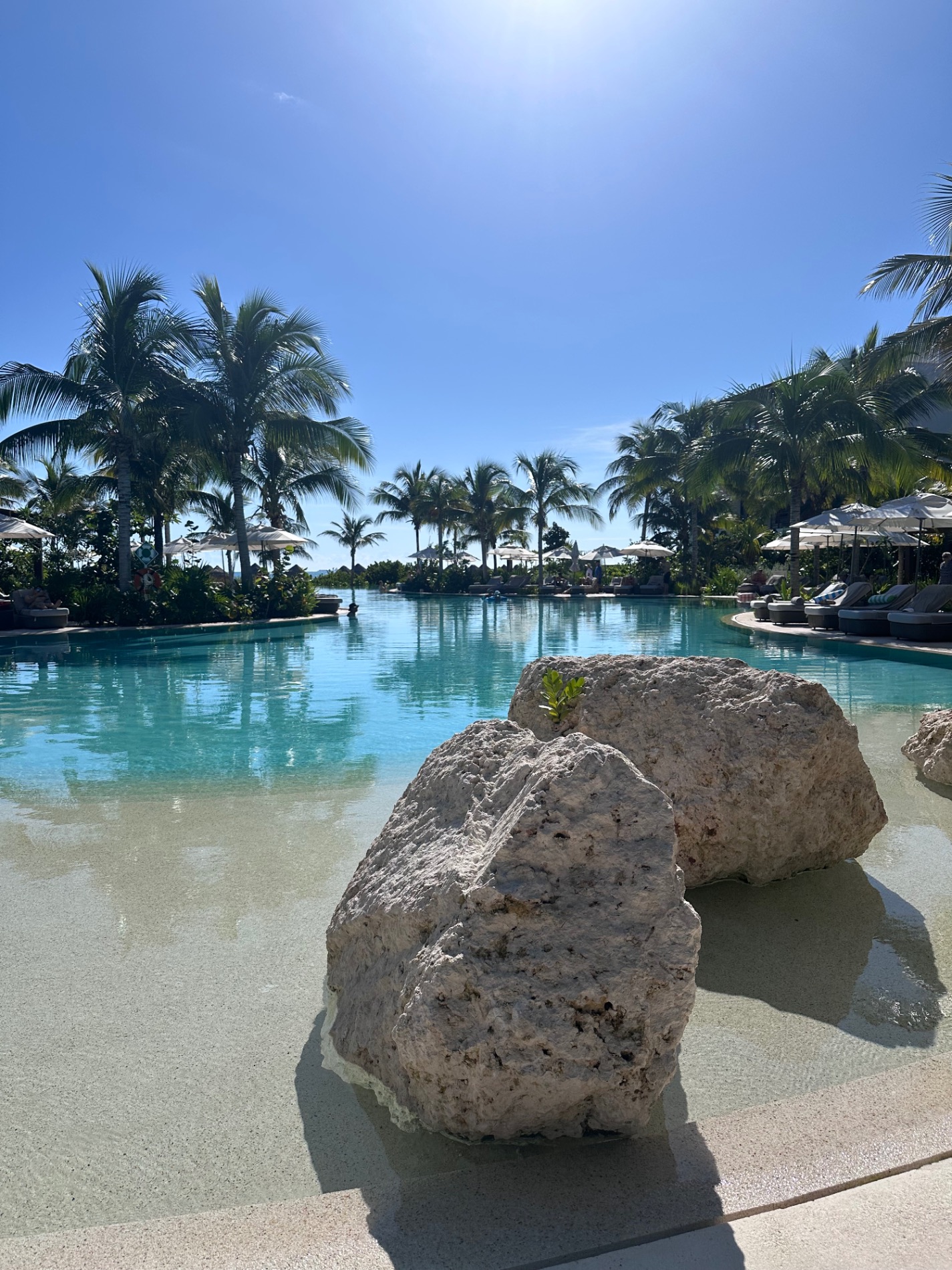 Turquoise lagoon with limestone rocks in shallow water, palm trees lining the shore, and resort loungers in background
