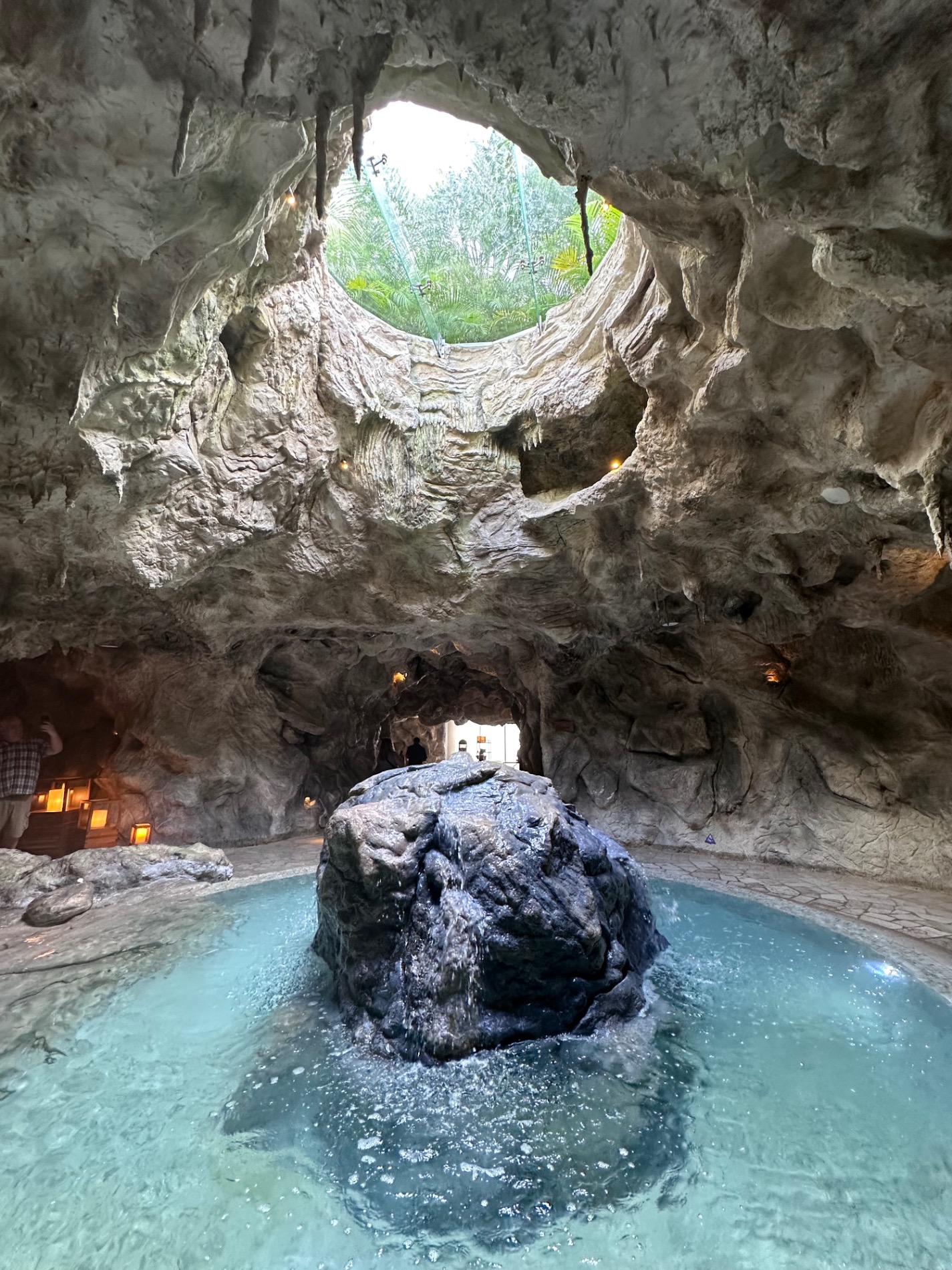 Natural cave with turquoise geothermal pool, limestone formations, and opening to tropical vegetation above