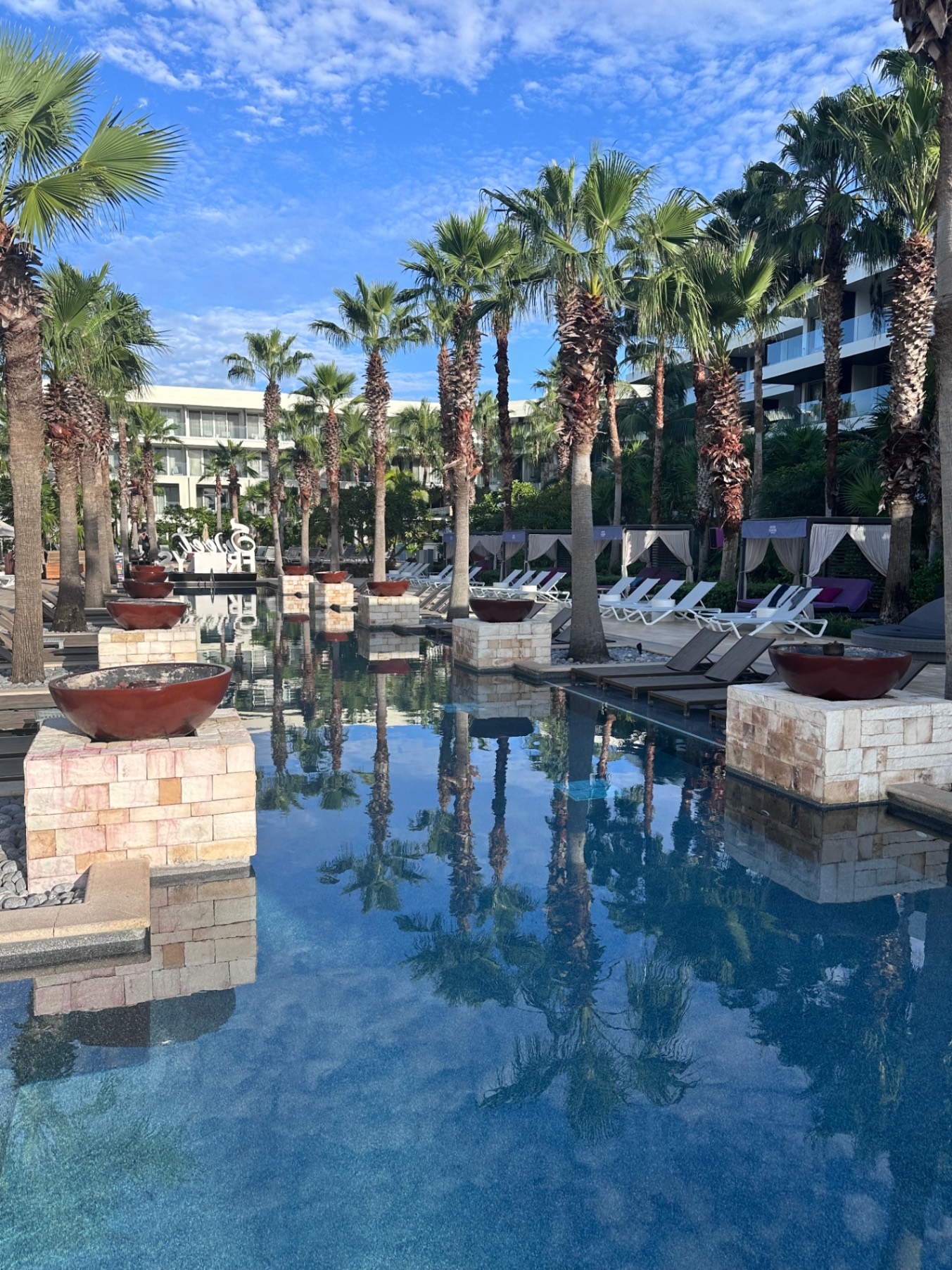 Modern resort courtyard with reflective water feature, palm trees, lounge chairs, and contemporary architecture