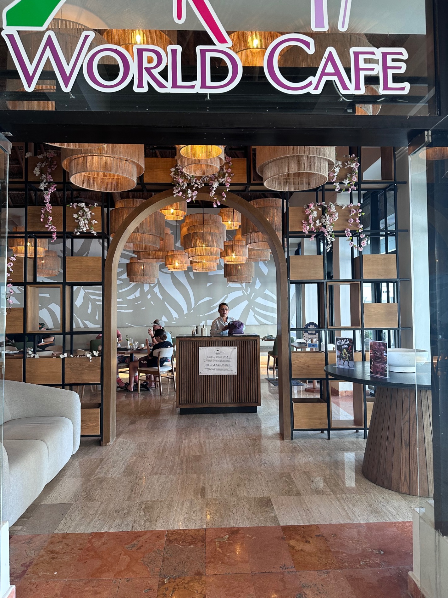 Modern café interior with neon 'World Cafe' sign, wooden pendant lights, floral installations, and contemporary design