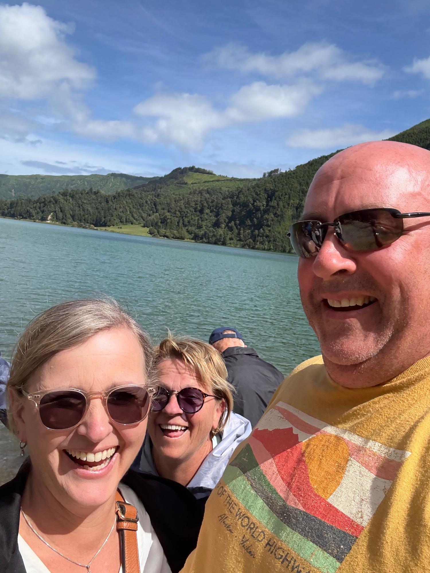 Three people smile at the camera while standing on a boat with turquoise water and green forested hills in the background