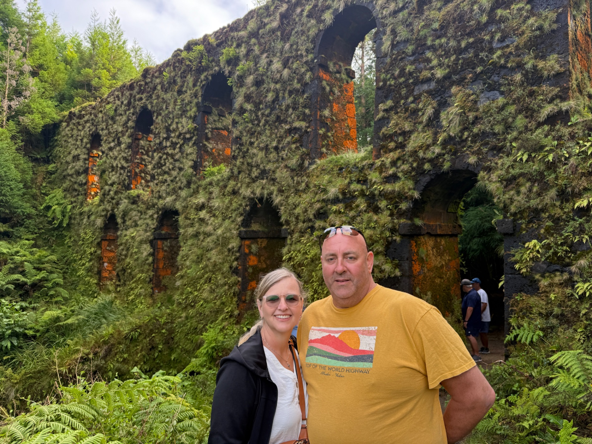Marcia White and a man stand in front of moss-covered stone ruins with arched openings in a lush green forest setting