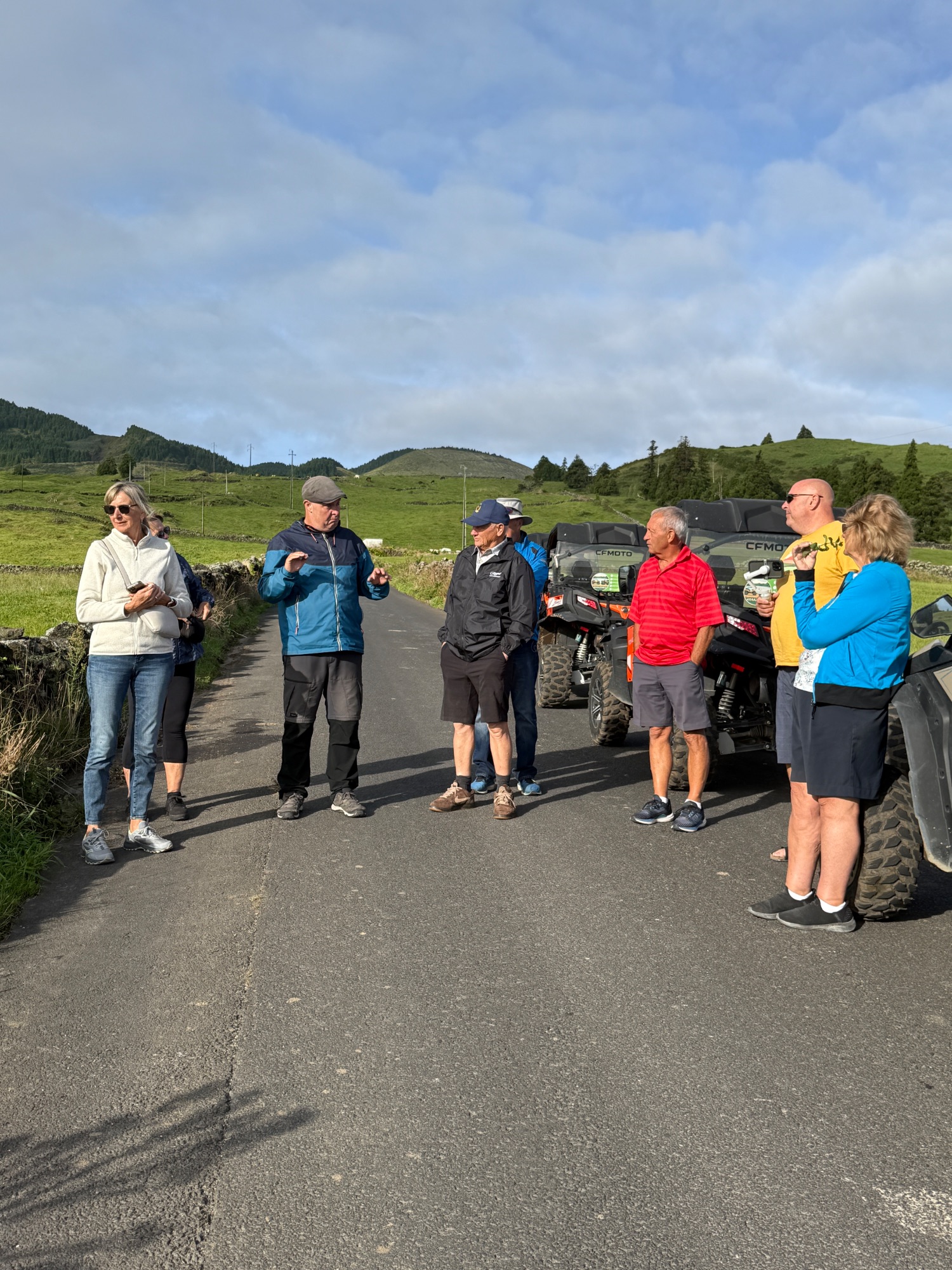 A group of tourists gathered on a rural road with all-terrain vehicles, surrounded by green rolling hills and mountains