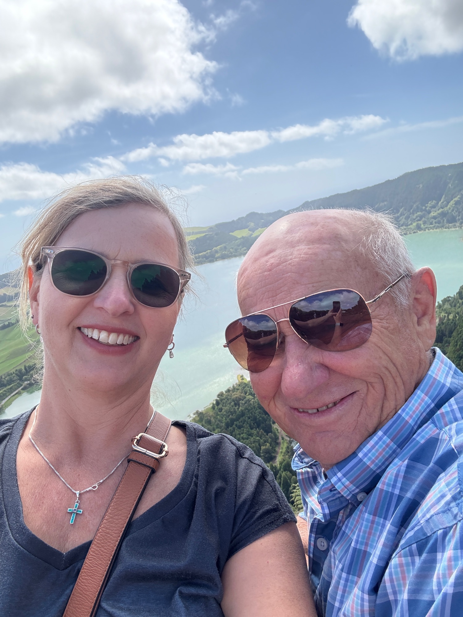 Marcia White and an older man smile at the camera with a turquoise lake and forested landscape visible behind them