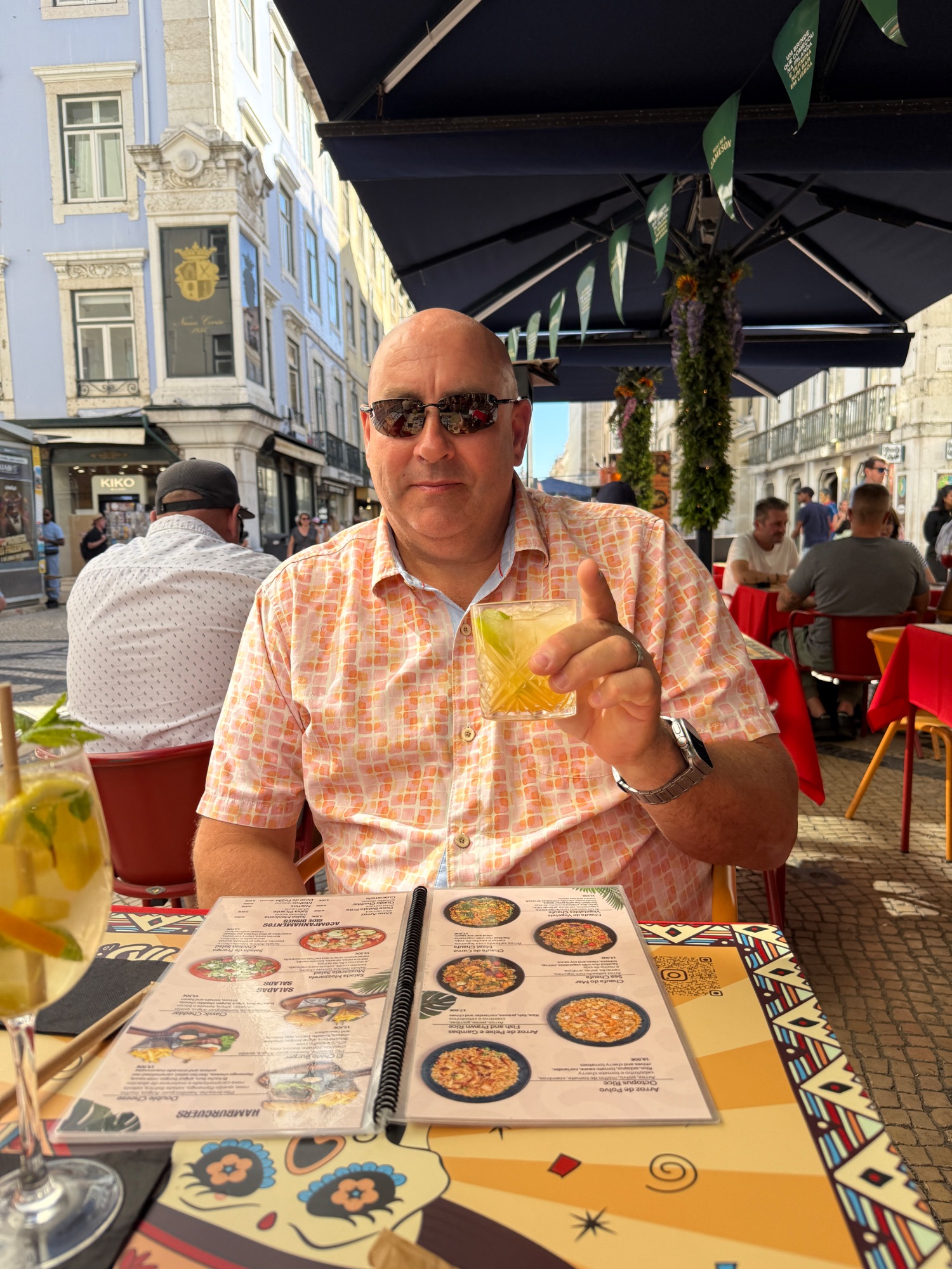 A man in an orange checkered shirt holds a drink while reviewing a menu at an outdoor café in a European town square