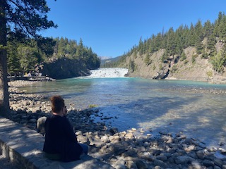 Person sitting on rocky shore beside a turquoise river flowing through forested valley with mountains