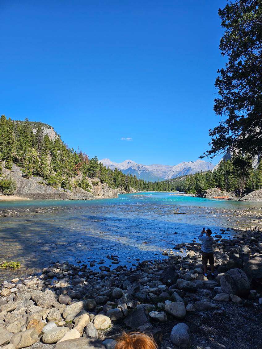 Turquoise river surrounded by rocky shores, coniferous forest, and snow-capped mountains under clear sky