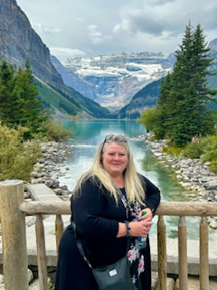 Blonde woman in dark jacket standing at wooden railing overlooking a turquoise alpine lake between mountains