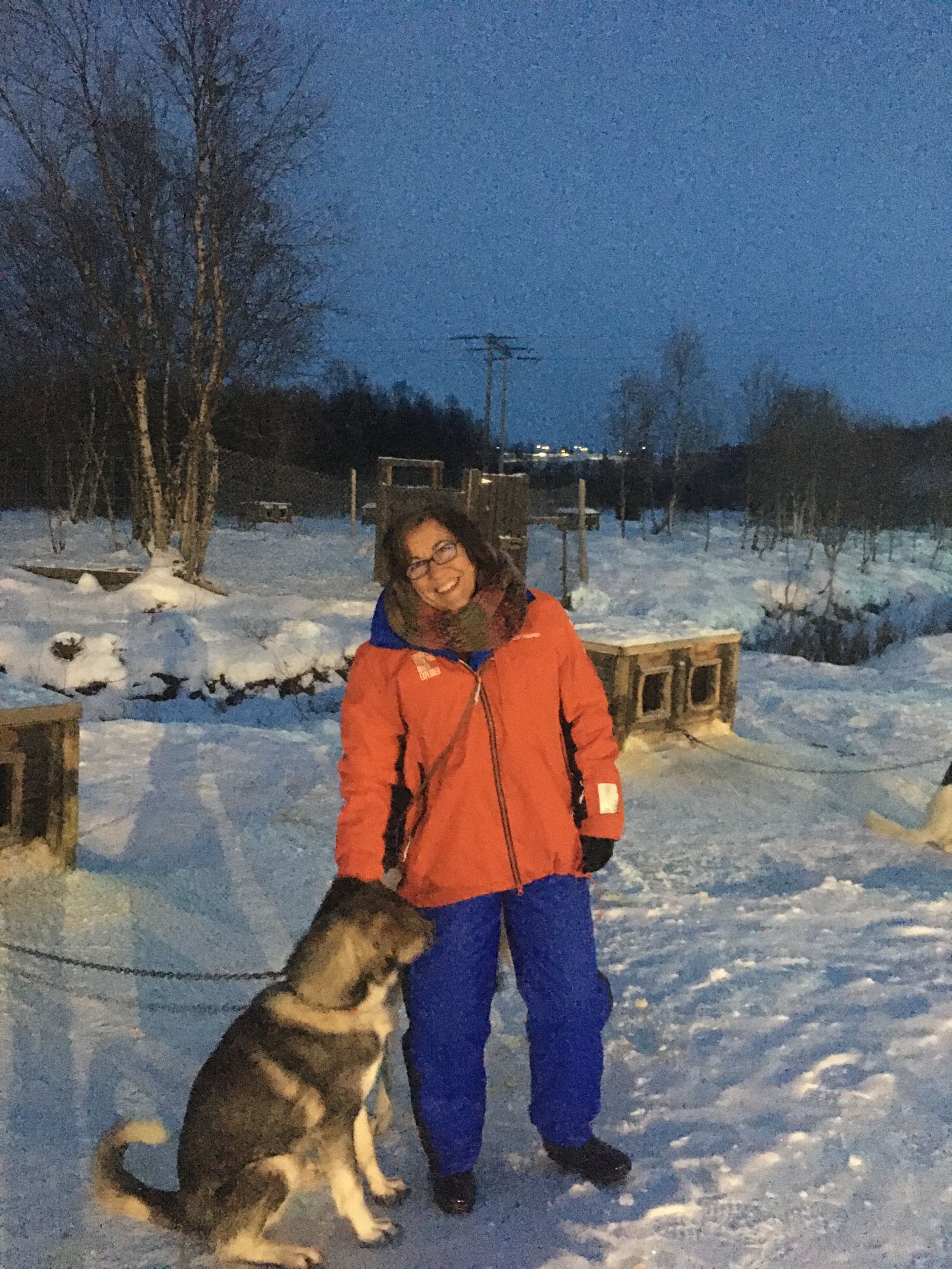 Woman in orange jacket smiling with husky dog in snowy landscape at dusk with wooden structures
