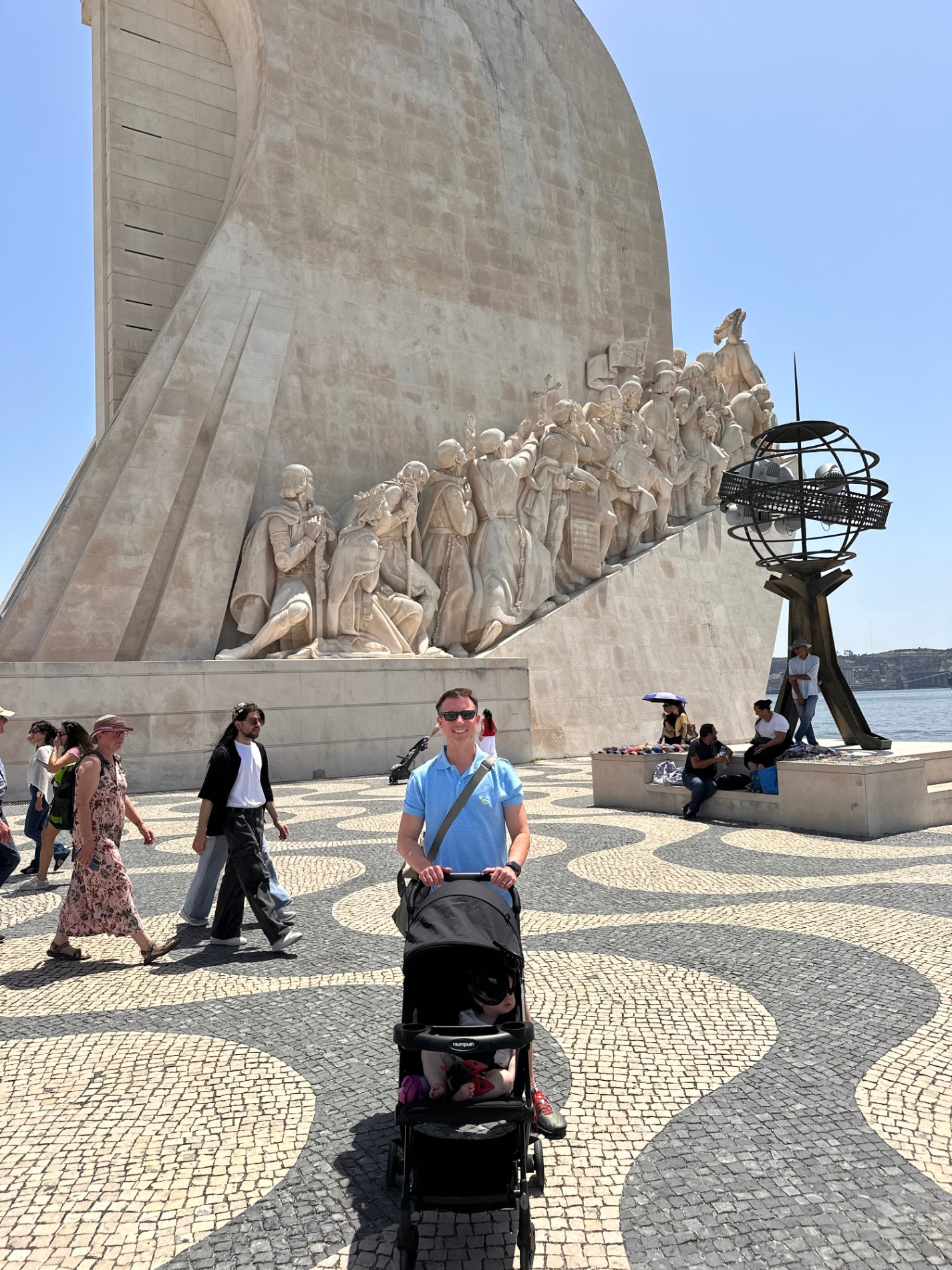 Man pushes a stroller past a large stone monument with carved figures at Lisbon's waterfront