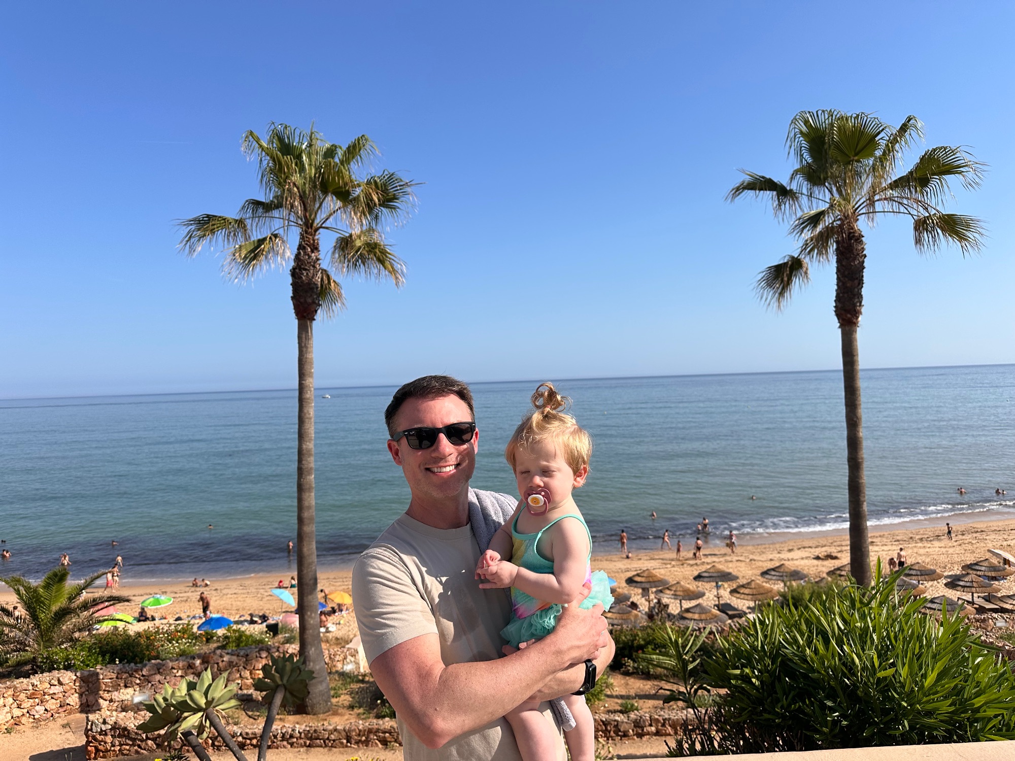 Man holds a toddler on a sandy beach with palm trees and calm turquoise water in the background