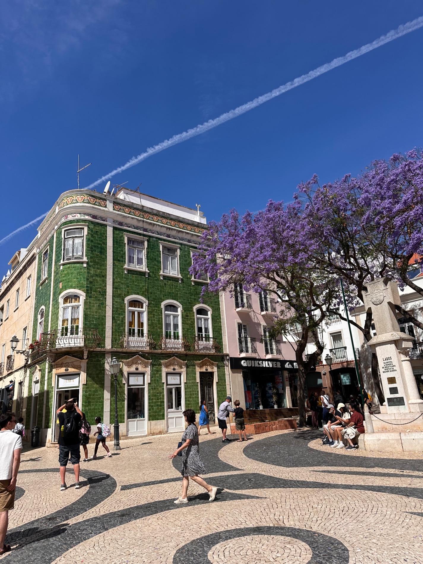 Green-tiled corner building with white trim and purple jacaranda flowers blooming nearby in Lisbon