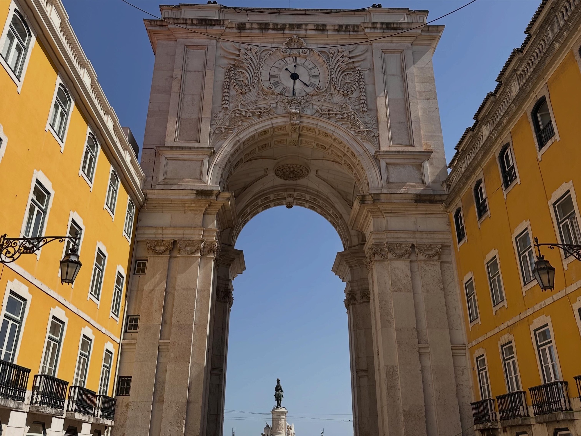 Ornate stone archway with clock tower framing a statue in a Lisbon plaza with yellow buildings