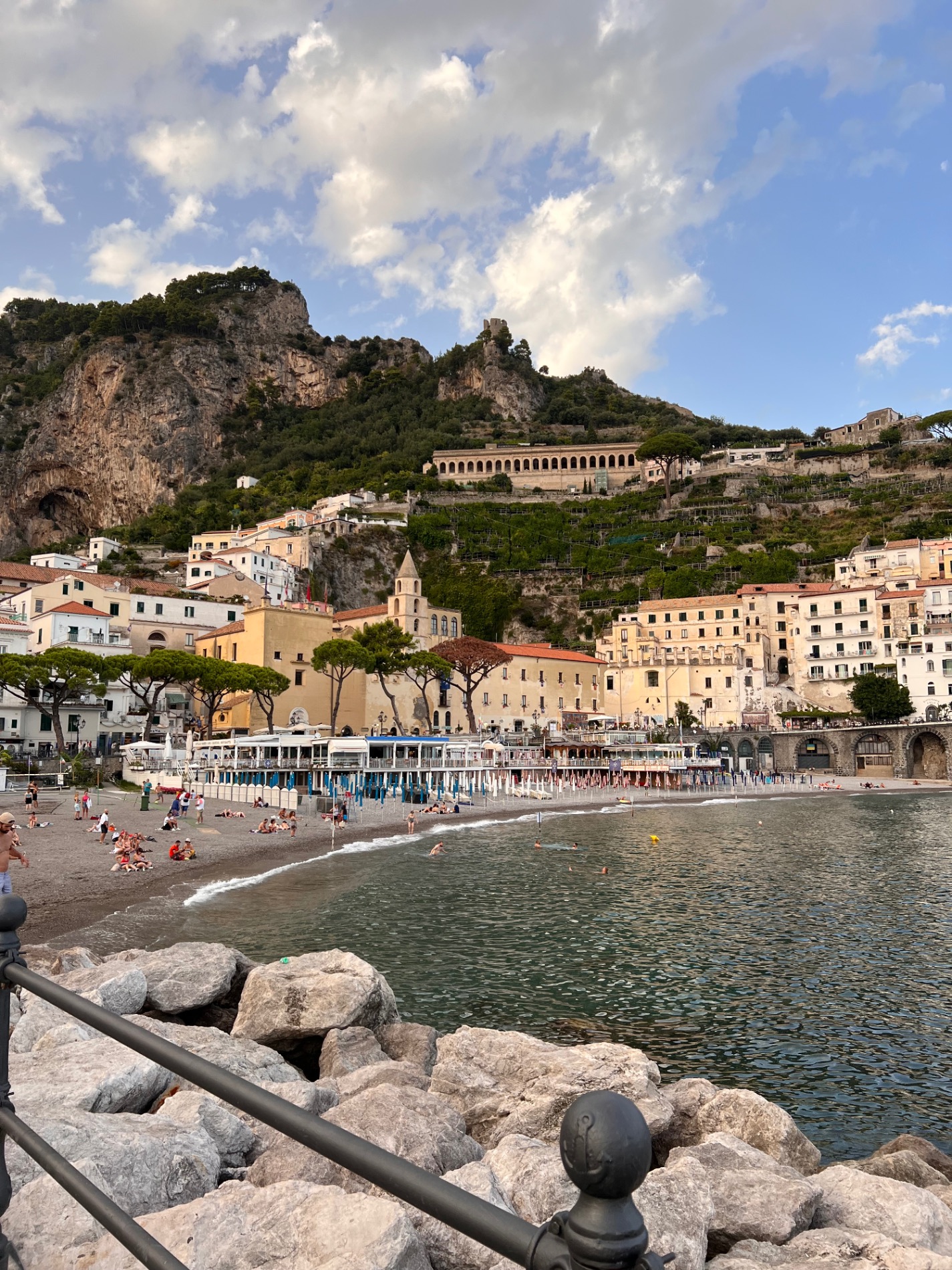 Colorful beachfront buildings nestled against steep cliffs with a sandy beach and swimmers in the turquoise Mediterranean Sea below