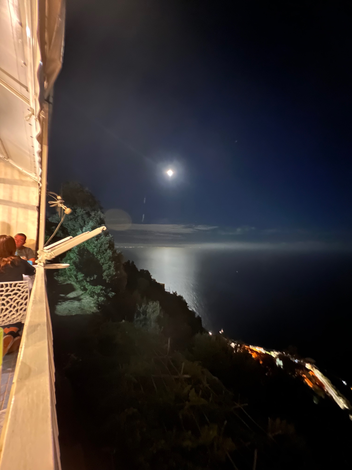 Moonlit seascape viewed from a clifftop terrace, with moonlight reflecting across dark water and distant coastal lights below