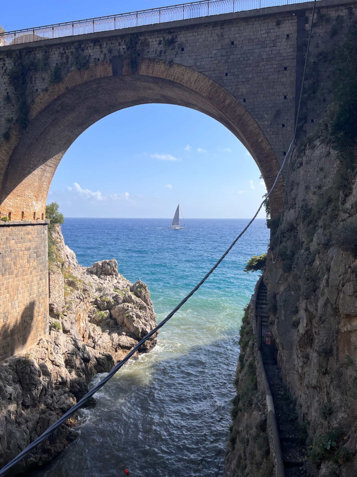 Sailboat visible through a large stone arch of a seaside fortification, with turquoise water and rocky coastline framing the view