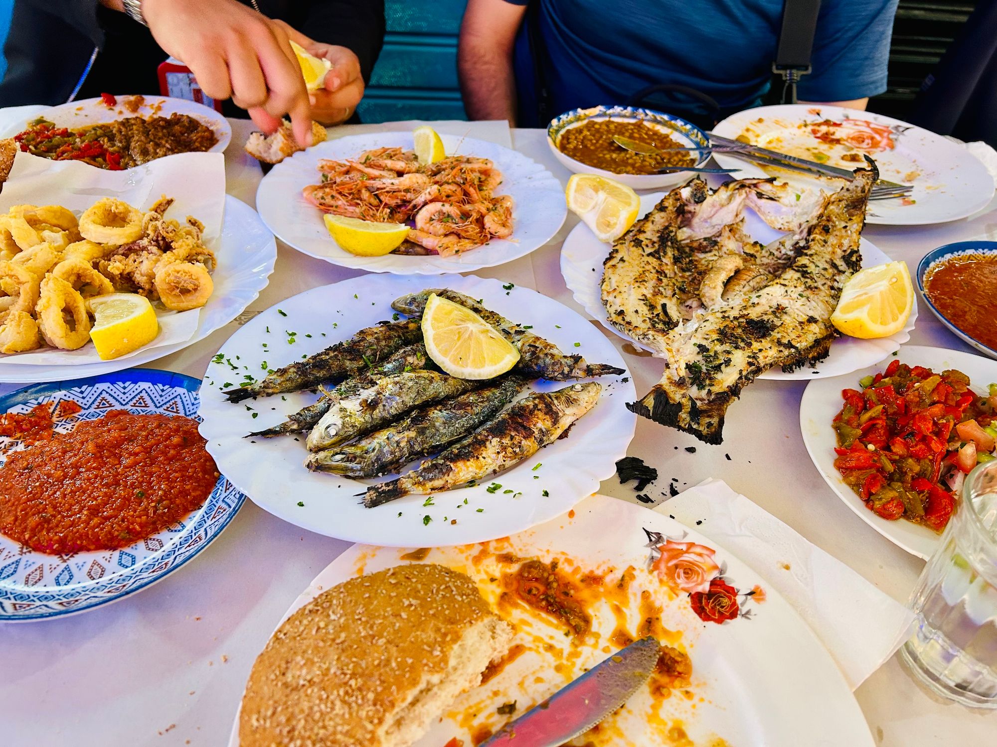 Spread of Moroccan seafood dishes including grilled fish, shrimp, calamari, and traditional mezze on white plates with lemon wedges