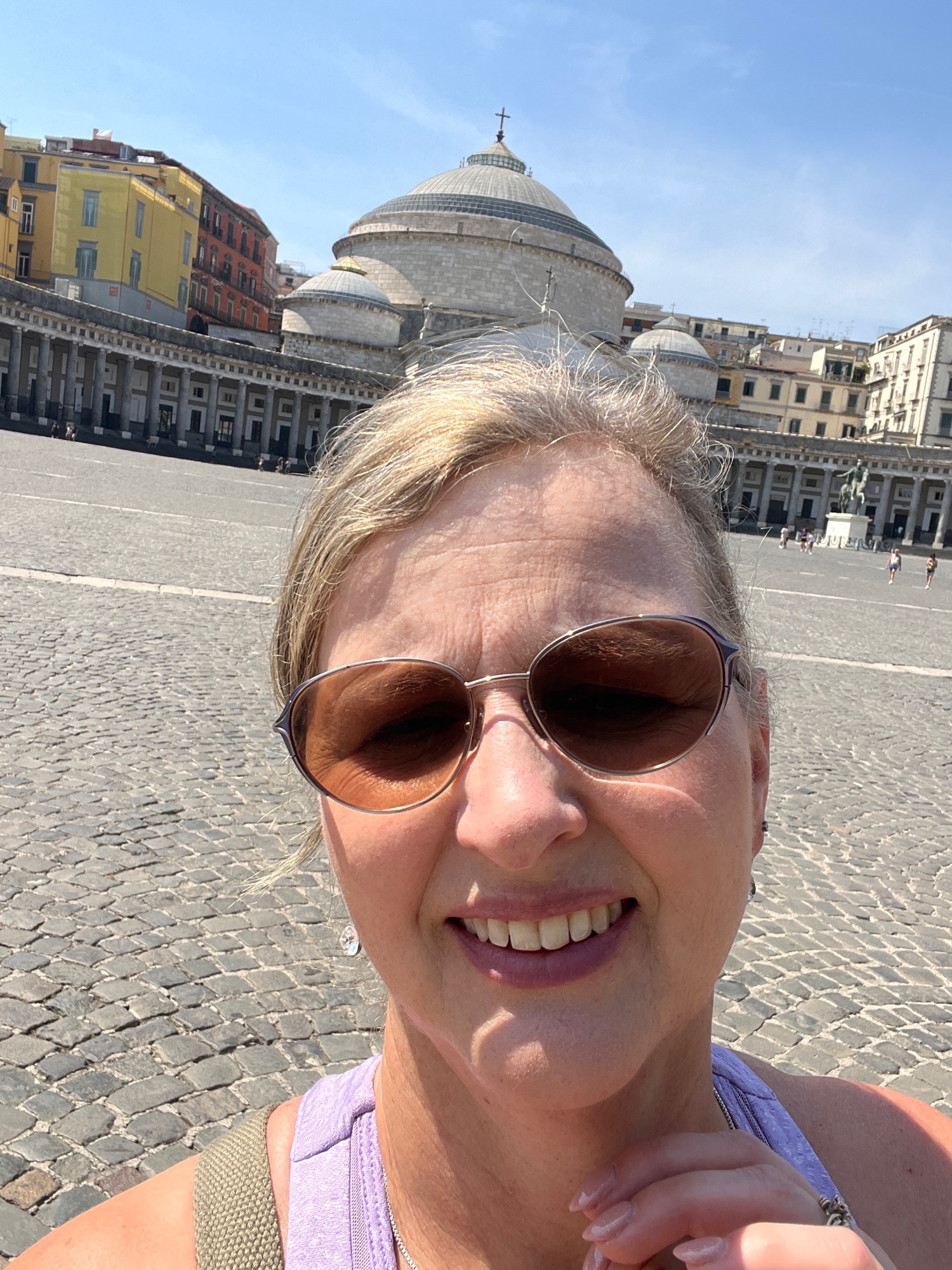 Woman in sunglasses smiling at Piazza del Plebiscito in Naples, with the dome of Basilica di San Francesco di Paola visible behind her