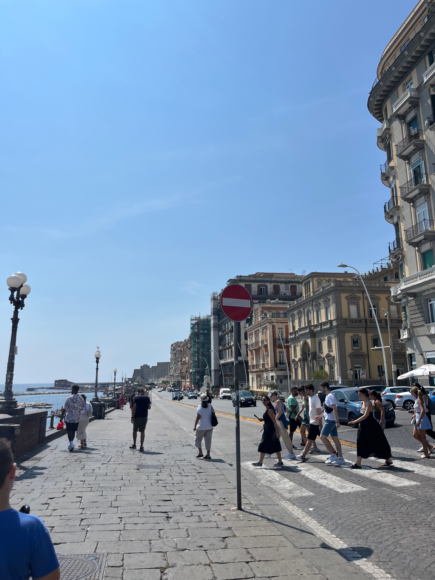 Waterfront promenade in Naples with colorful historic buildings, pedestrians, and a red traffic sign on a sunny day