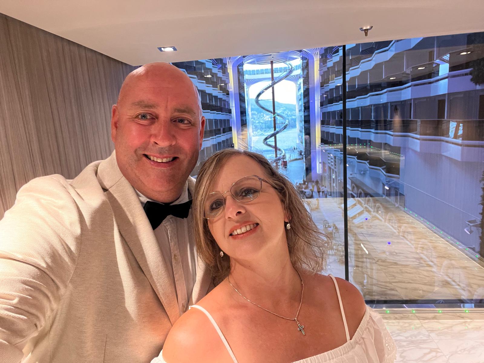 Couple in formal attire posing in a modern hotel room with floor-to-ceiling windows overlooking a Mediterranean coastal city