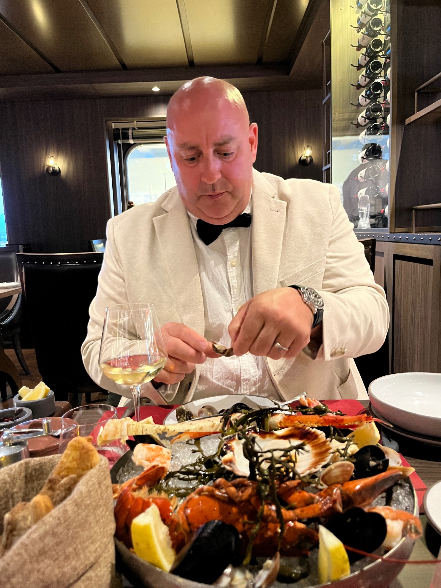 Man in a cream blazer and bow tie enjoying seafood at an elegant dinner table with wine and lobster