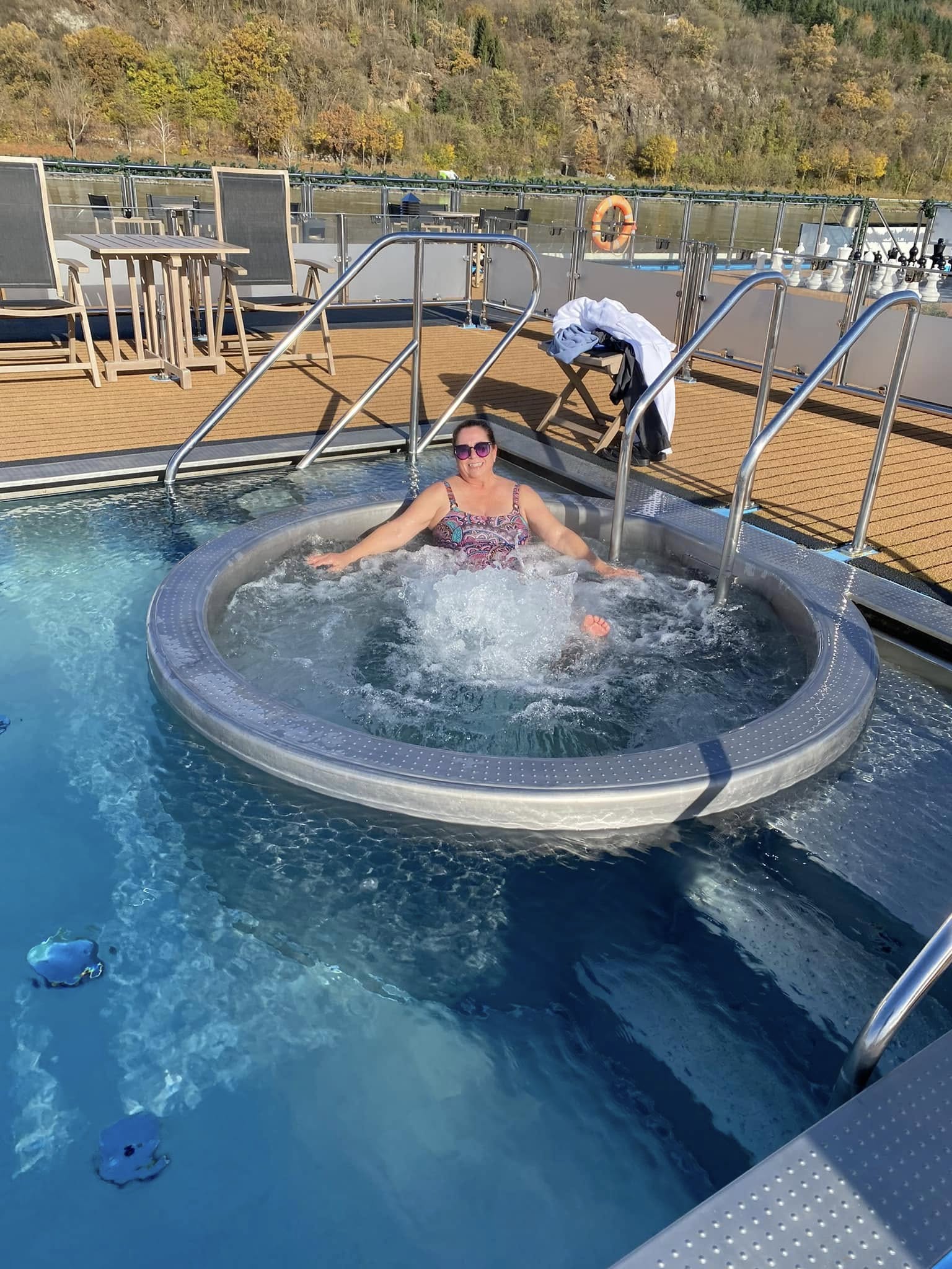 Woman in a swimsuit relaxing in a circular hot tub on a cruise ship deck with forest-covered hills visible in the background