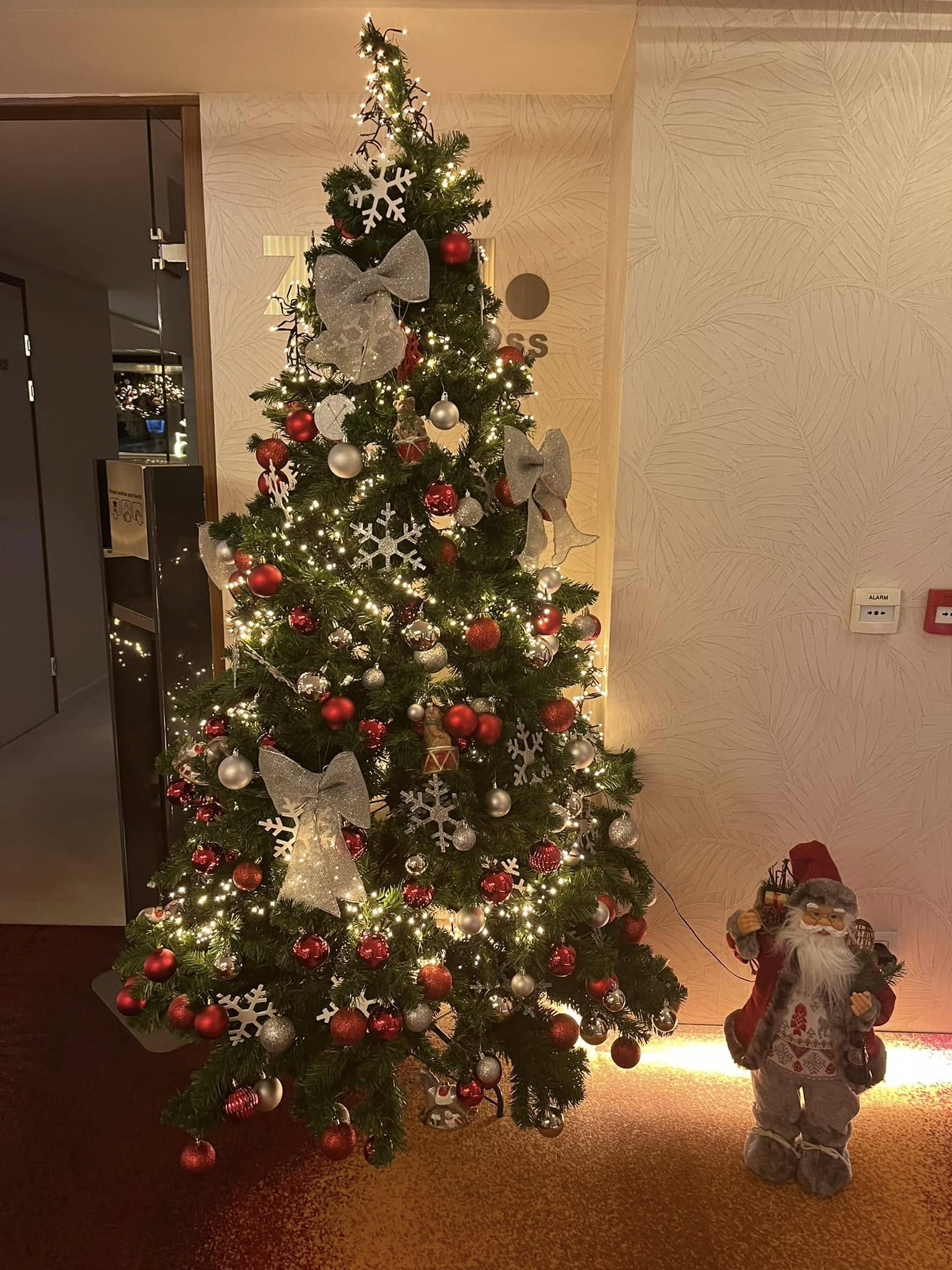 Christmas tree decorated with red and gold ornaments, silver bows, snowflake decorations, and white lights next to a Santa figurine