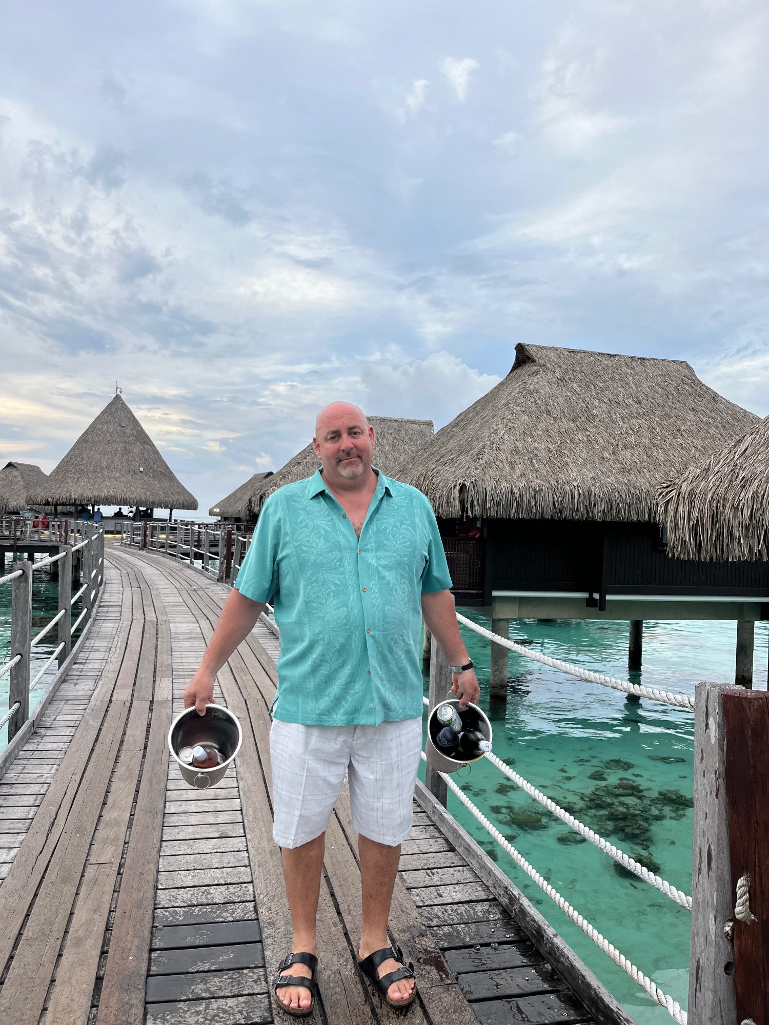 Man in turquoise shirt holding two black buckets on a wooden dock with overwater bungalows and turquoise lagoon