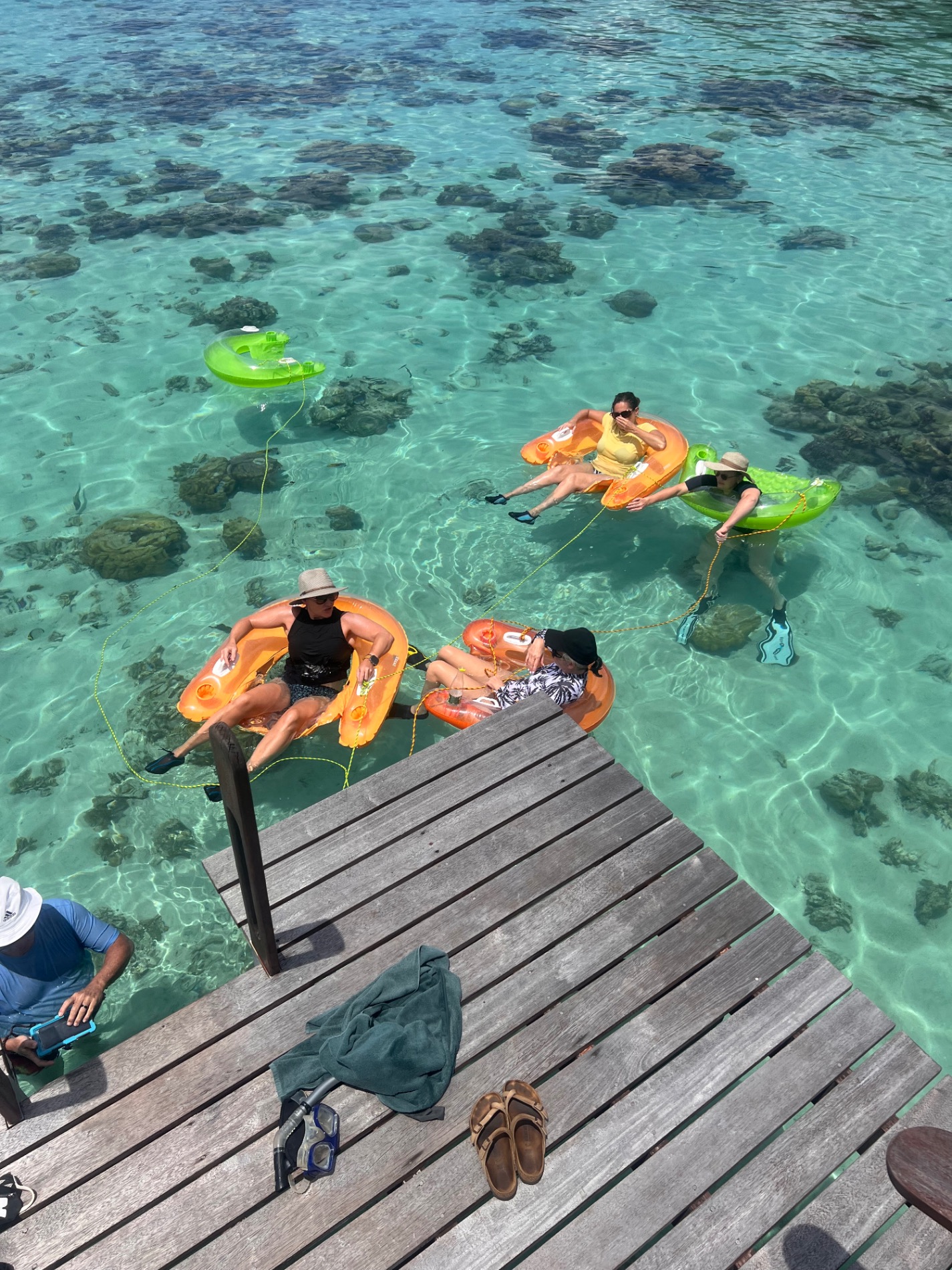 Four people on colorful floats in crystal-clear shallow water with coral visible below