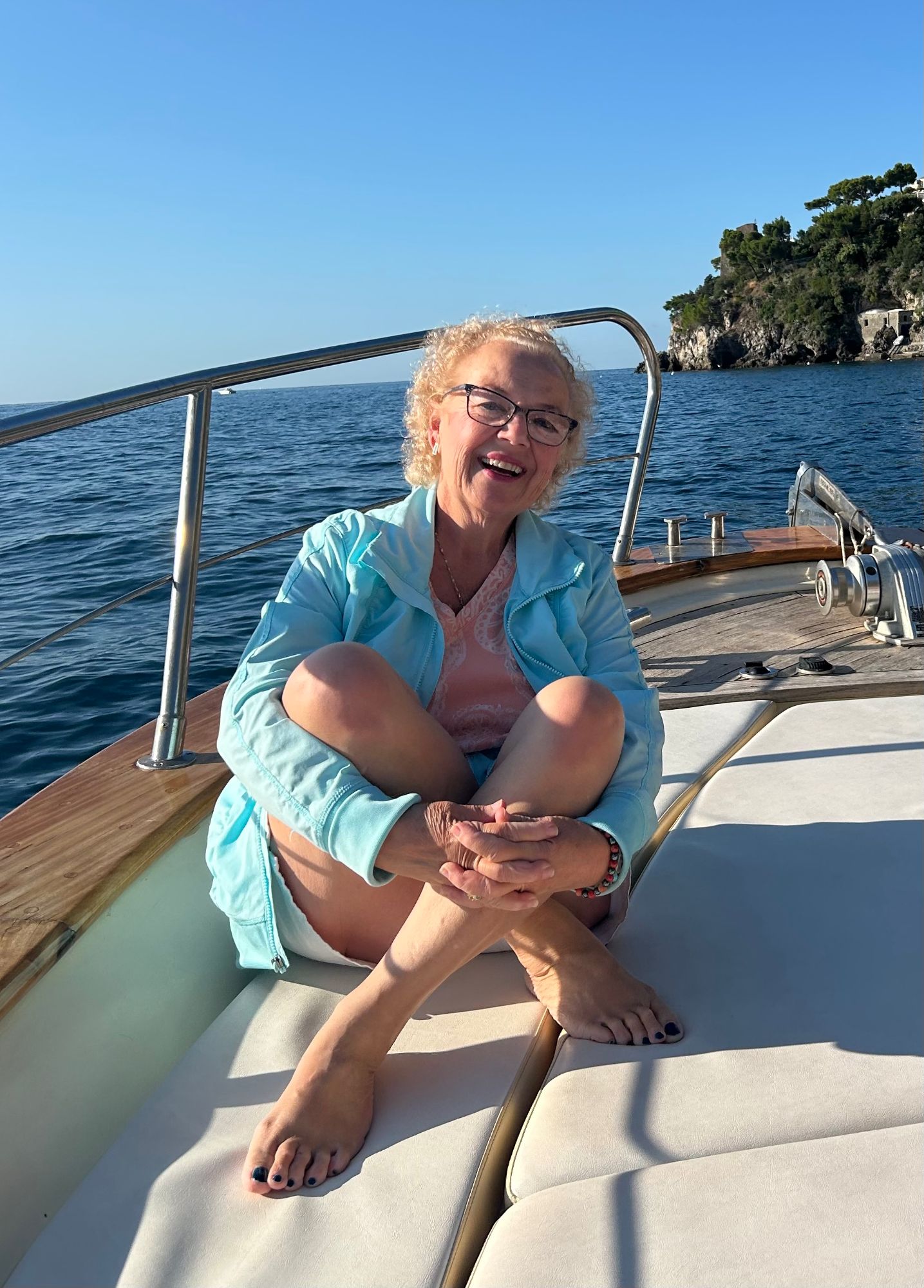 Patti Carter sitting on a boat deck laughing, wearing a light blue jacket with ocean and rocky coastline behind her