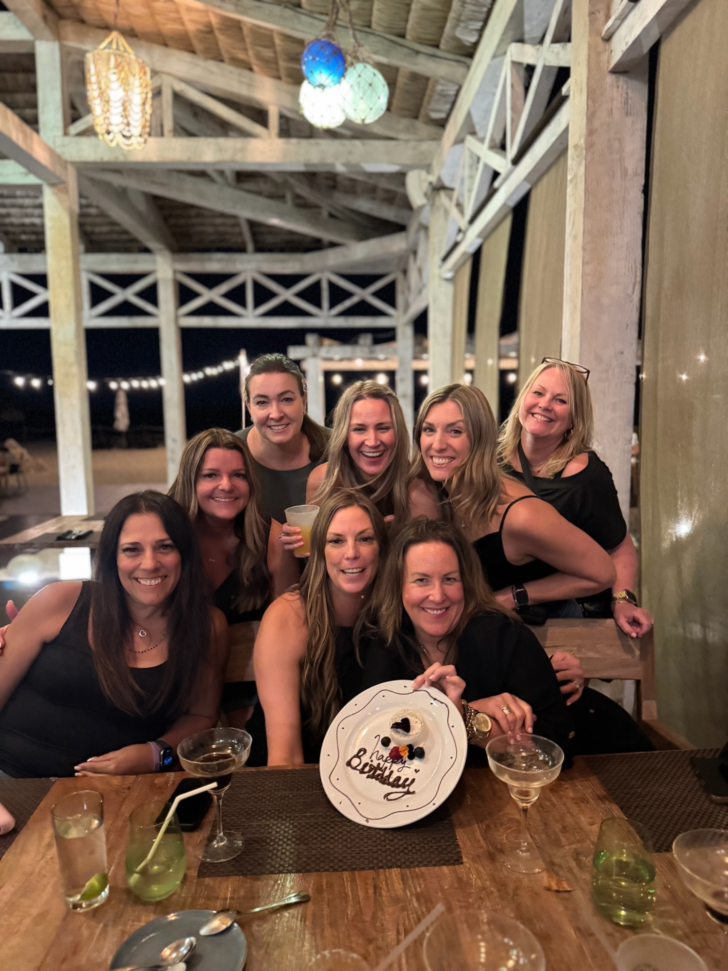 Group of women gathered around a table with a birthday cake at an outdoor beachside venue at night