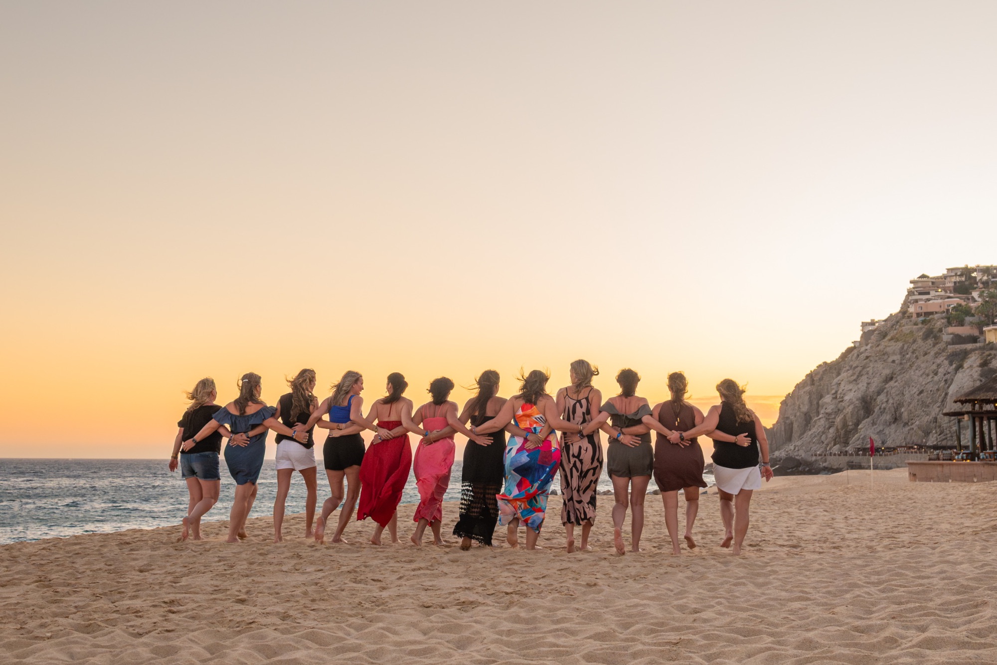Line of twelve women standing on a beach at golden hour with dramatic cliff formations and ocean in the background