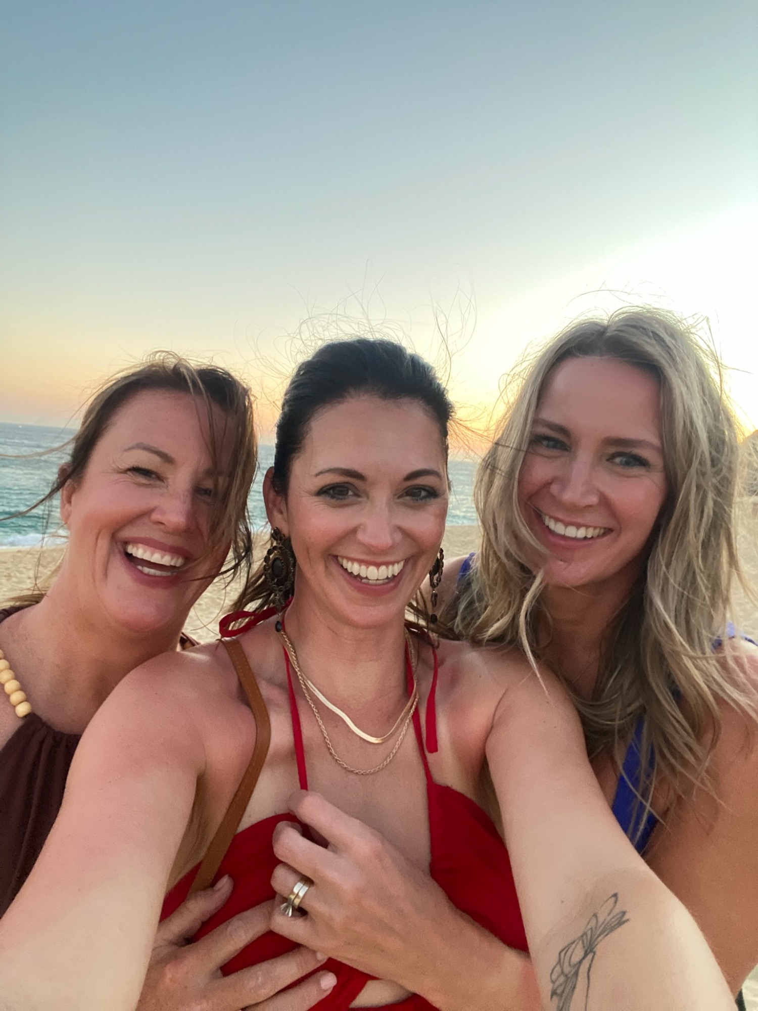 Three women smiling together for a selfie on a beach at sunset in Cabo San Lucas
