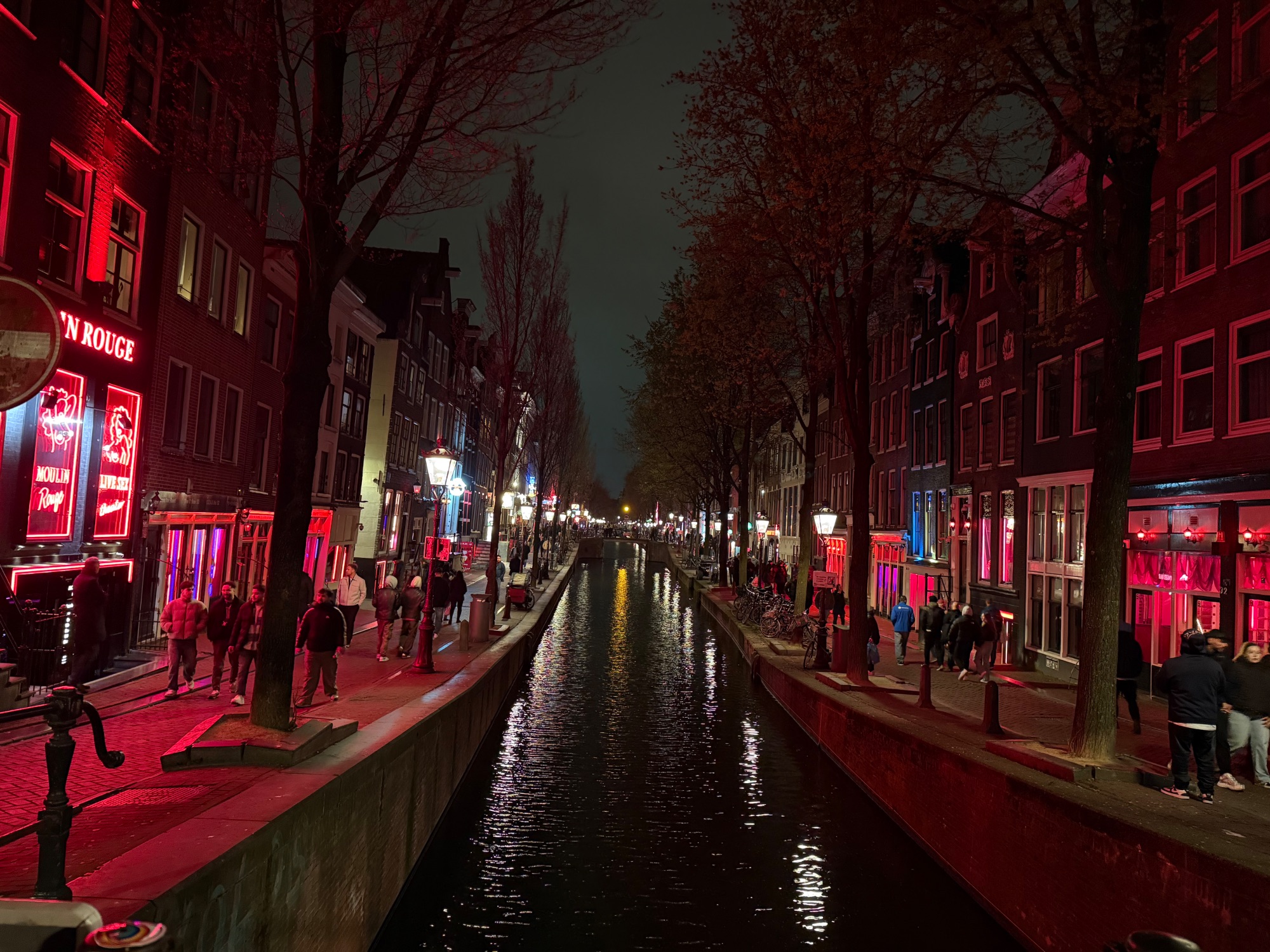 Red neon-lit street in Amsterdam's Red Light District at night, with historic buildings reflected in a canal
