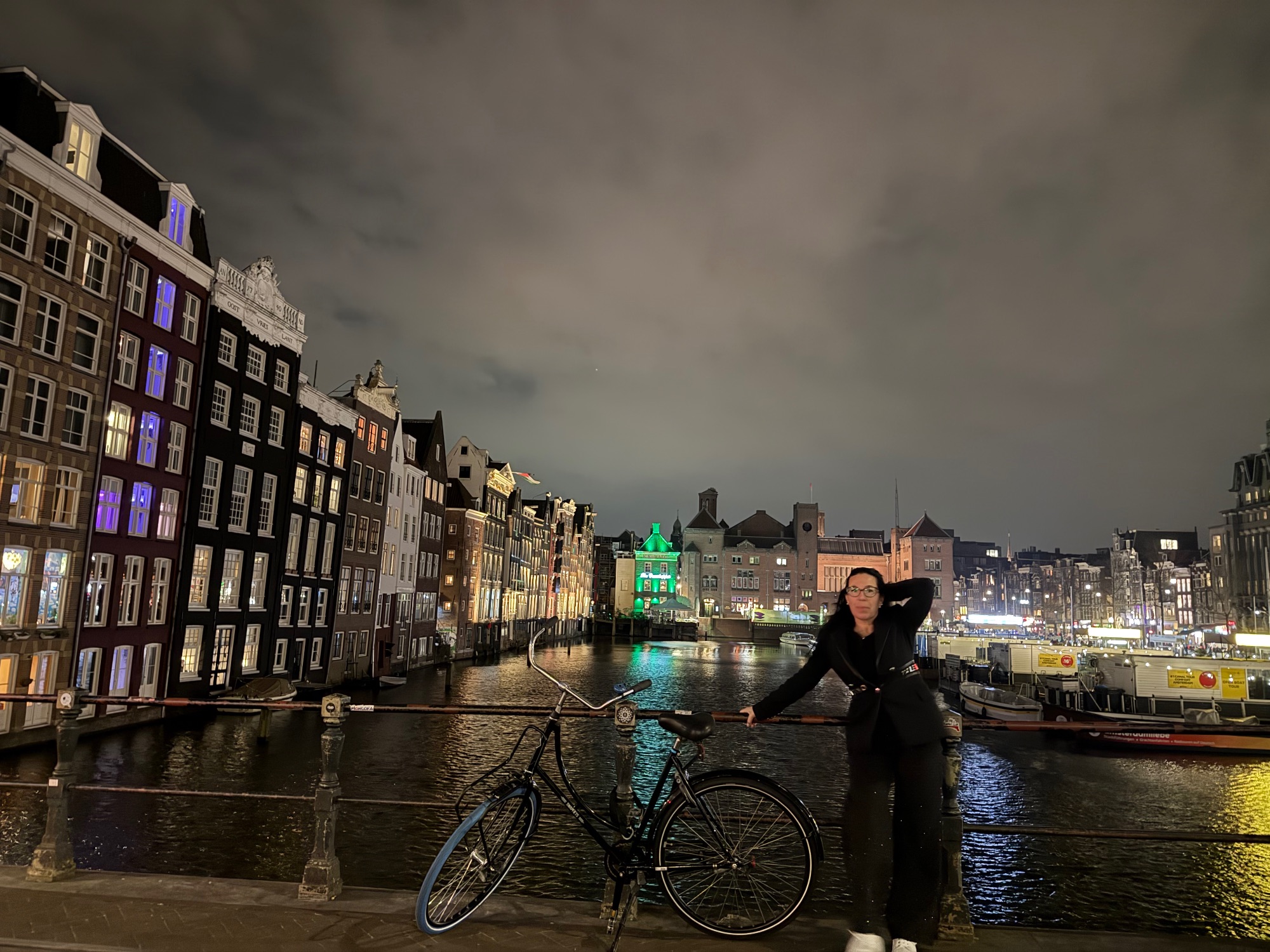 Woman posing with a bicycle by a canal in Amsterdam, with historic buildings and green-lit architecture behind her