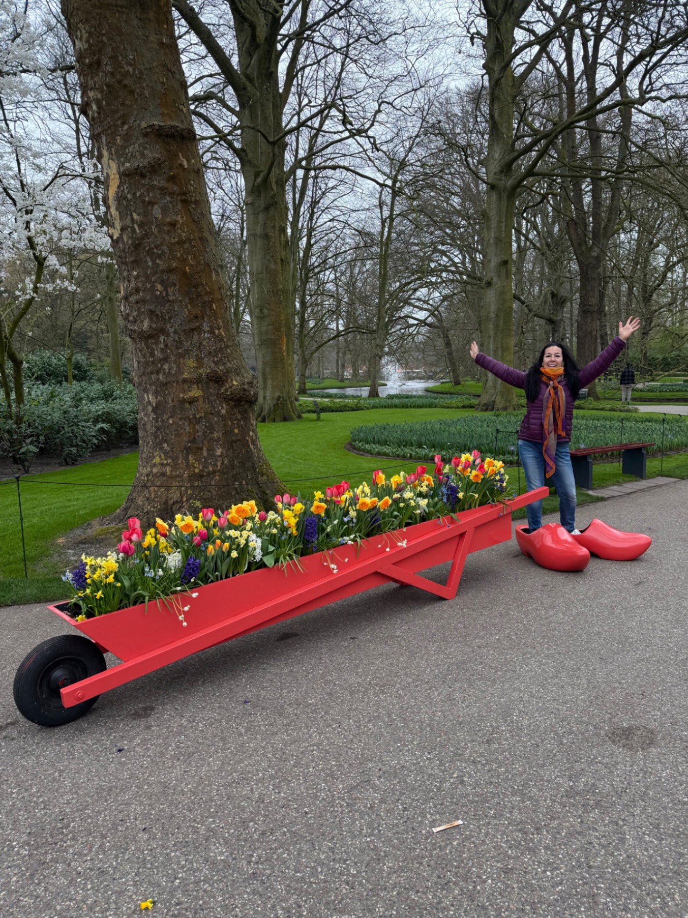 Woman standing on a red flower-filled seesaw sculpture in a park with bare trees and a pond