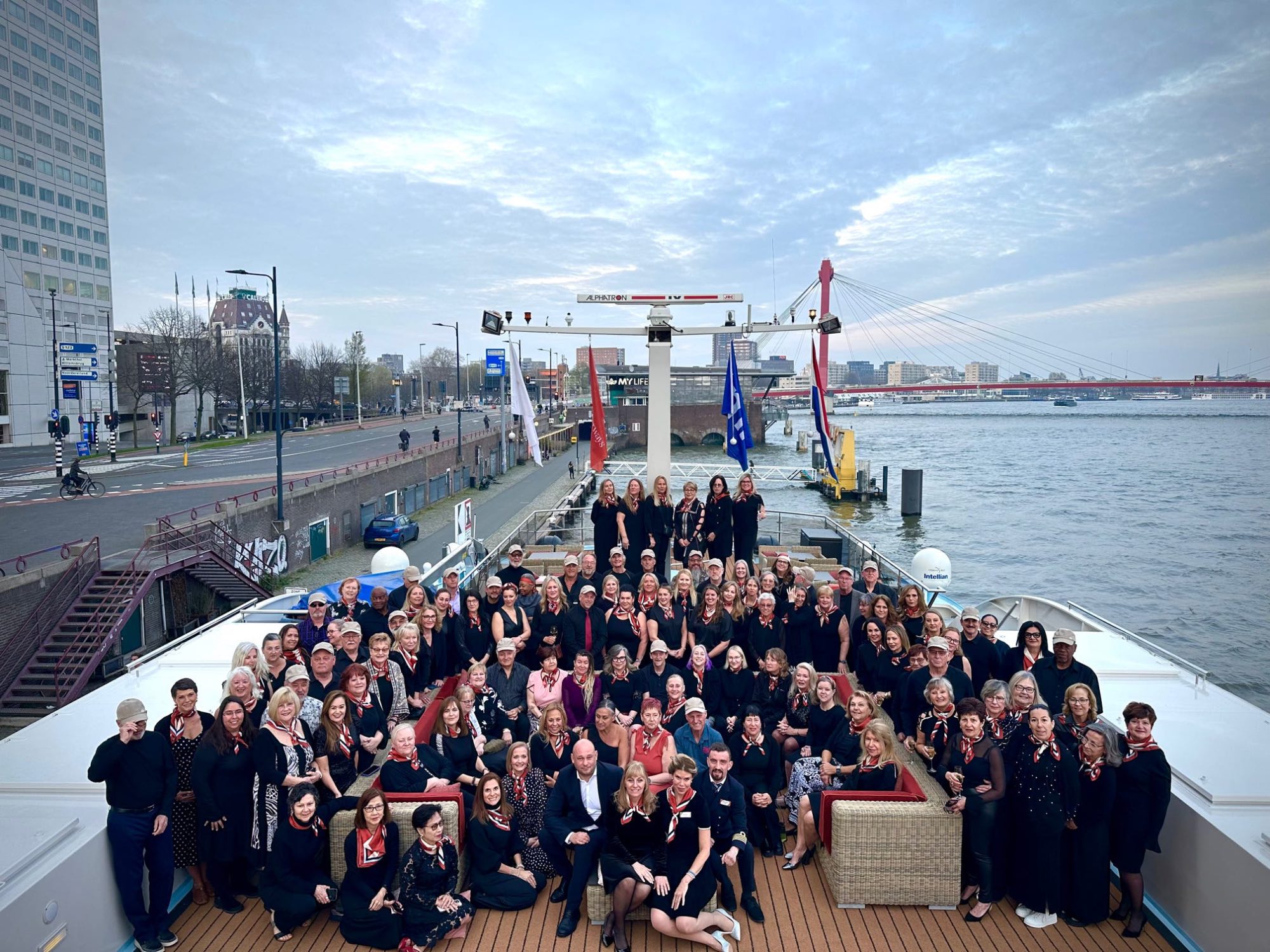 Large group of people gathered on a river cruise boat deck with Amsterdam's skyline and bridges visible
