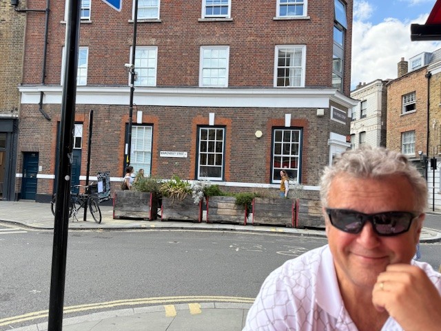 A man in sunglasses and light pink shirt poses on a London street in front of a brick building