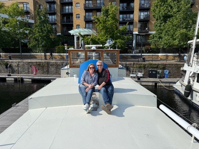 A couple sits on the bow of a white boat moored at a London canal with modern apartment buildings visible