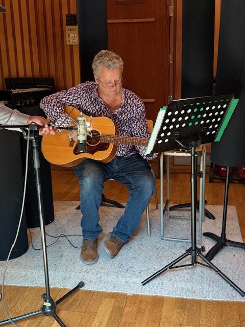 A man plays an acoustic guitar while reading sheet music from a stand in a wooden-floored studio space