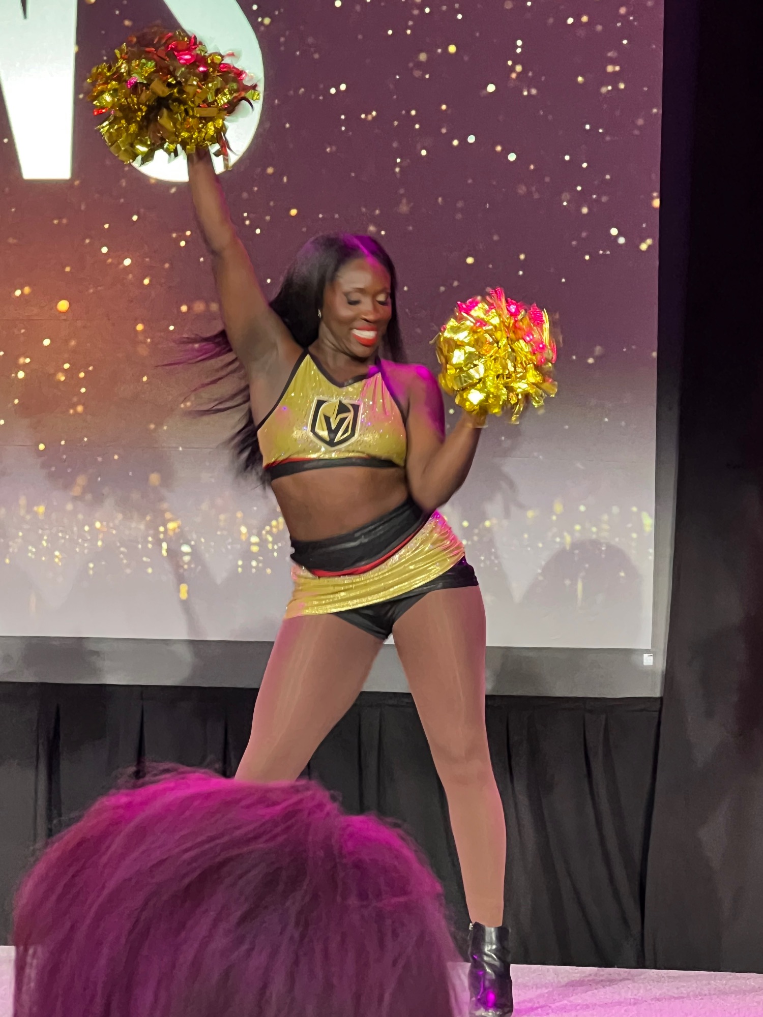 Cheerleader in gold and black Vegas Golden Knights uniform holding pom-poms on stage with sparkly backdrop