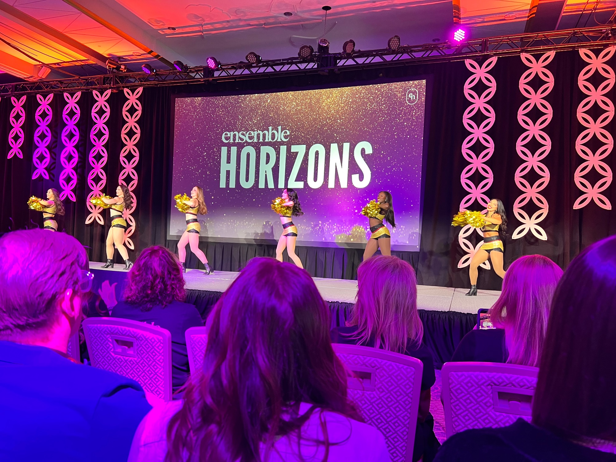 Ensemble Horizons event stage with performers in gold outfits and pink geometric backdrop panels under purple lighting