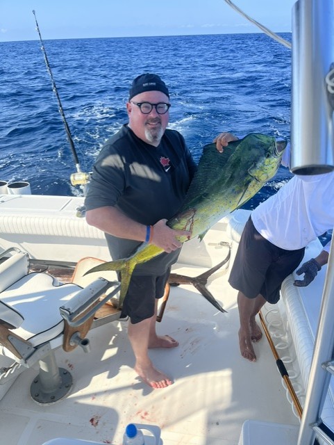 Man in black shirt and cap holds a large yellow-green mahi-mahi fish on a fishing boat