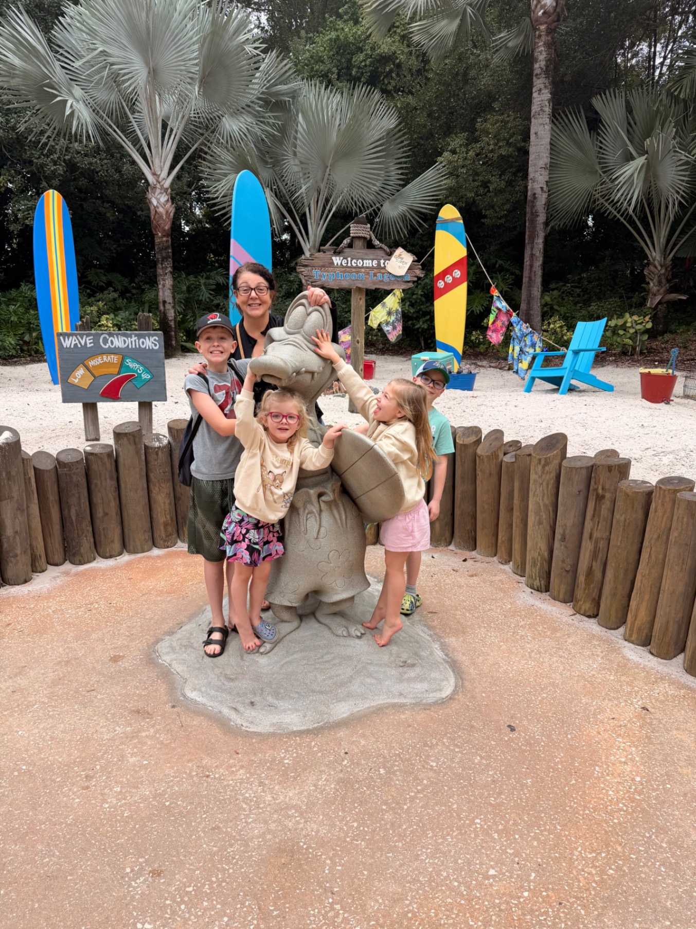 Four children and two adults posing with a large sand sculpture of a hippo at a beach-themed park with palm trees